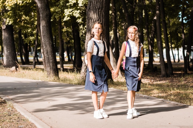 Dos niñas de la escuela primaria sonríen y se toman de la mano y van a la escuela en un día cálido