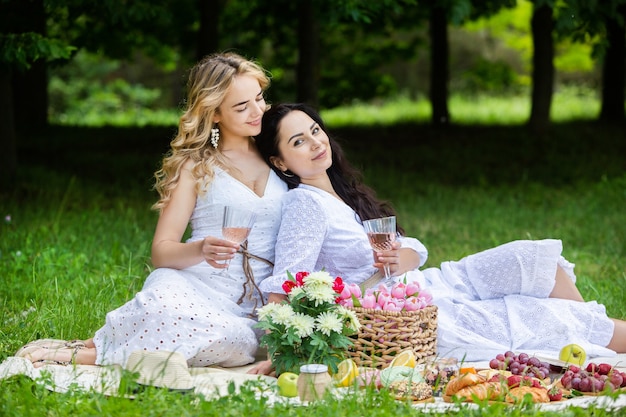 Dos niñas están descansando en el parque sentadas en una manta de picnic con frutas y vino