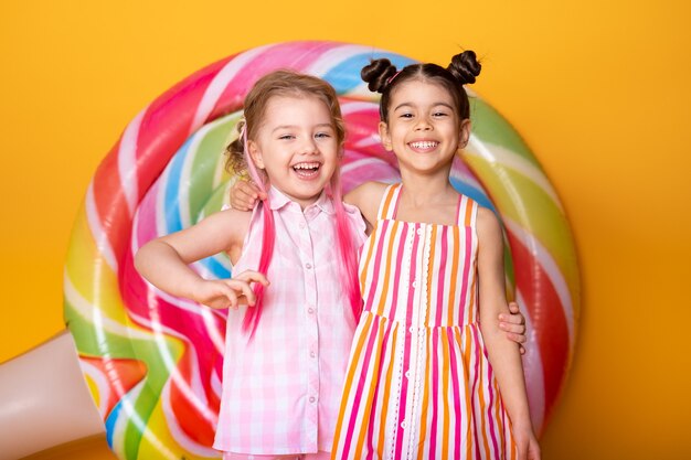 Dos niñas felices en vestido colorido riendo abrazándose divirtiéndose con piruleta.