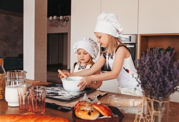 Dos niñas hacen masa juntos en la cocina. Las hermanas juegan con sombreros blancos del jefe.