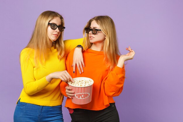 Dos niñas de hermanas gemelas rubias jóvenes irritadas en gafas imax 3d viendo películas, sostienen palomitas de maíz aisladas en la pared azul violeta pastel. Concepto de estilo de vida familiar de personas.