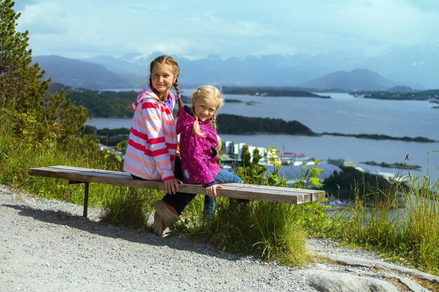 Dos niñas hermanas sentadas en un banco en la ciudad de Alesund y las montañas hasta el fiordo en el fondo, Noruega