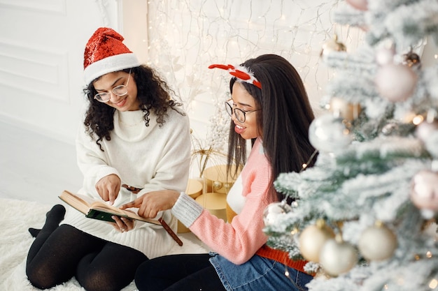 Dos niñas indias con anteojos sentadas cerca del árbol de Navidad y leyendo un libro