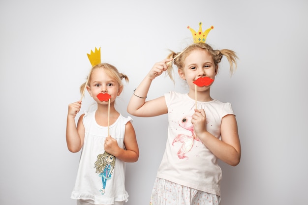 Dos niñas lindas con corona de papel y labios rojos posando fondo blanco en casa