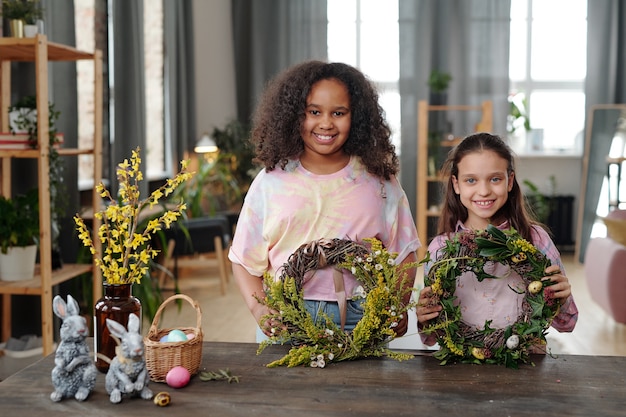 Dos niñas lindas felices en ropa casual mirándote con sonrisas mientras están de pie junto a la mesa de madera en el hogar y mostrando coronas de flores