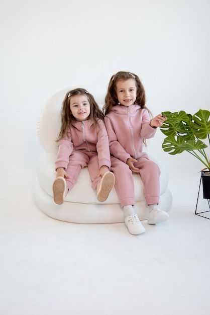 Dos niñas lindas con un mono rosa se sientan en una silla blanca Fondo blanco Infancia de moda