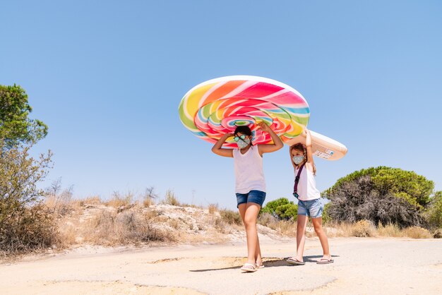 Dos niñas con una máscara en la cara y gafas caminando por un camping de pino con una colorida rueda de colchón inflable en la cabeza para disfrutar de unas vacaciones en medio del covid 19