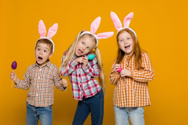 Dos niñas y un niño con orejas de conejo de Pascua sosteniendo huevos coloridos