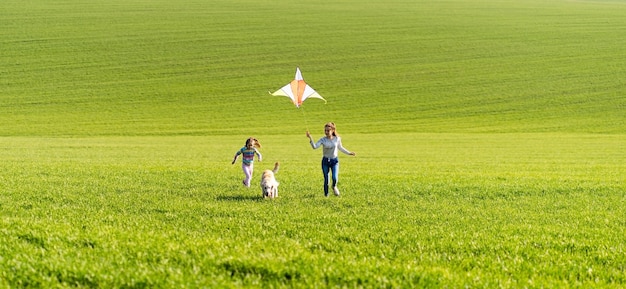 Dos niñas y un perro corriendo en el campo verde y sosteniendo una cometa voladora