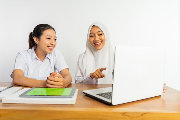 Dos niñas de secundaria sentadas en un escritorio con tableta y libros mientras usan una computadora portátil en un fondo aislado