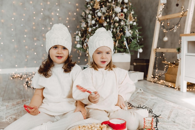 Dos niñas con sombreros de punto de invierno están sentadas en el suelo cerca del árbol de Navidad y comen palomitas de maíz. concepto de navidad.