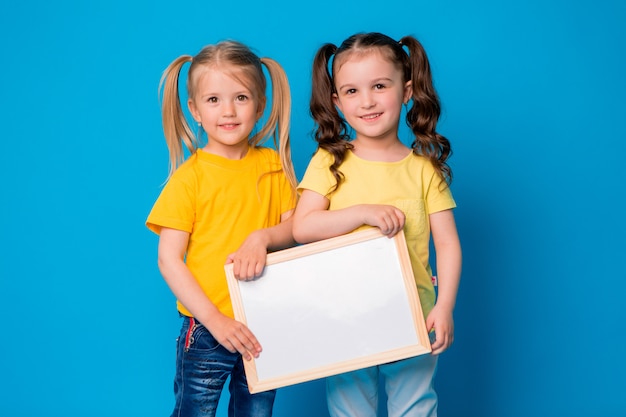 Dos niñas sonriendo con un tablero de dibujo vacío sobre un fondo azul.