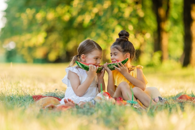 Dos niñas sonrientes jugando a sentarse para hacer un picnic y comer un trozo de fruta de sandía en el exterior