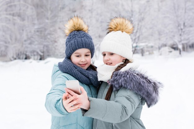 Dos niñas sonrientes con ropa de abrigo se toman una selfie en un teléfono inteligente en un parque de invierno cubierto de nieve Uso de la tecnología en el estilo de vida