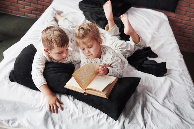 Dos niños se acuestan en una cama grande y leen un libro interesante. Están vestidos con el mismo pijama