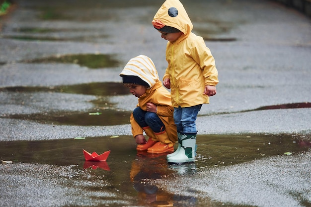 Dos niños con capas impermeables amarillas y botas jugando con un bote de papel hecho a mano al aire libre después de la lluvia juntos