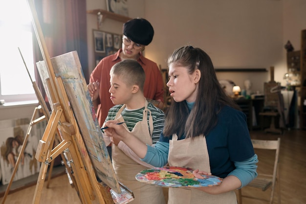 Foto dos niños con discapacidad pintando en caballetes en la clase de arte y el maestro