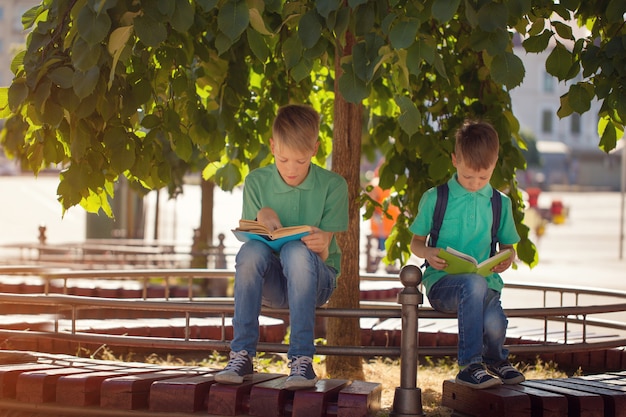 Dos niños escolares sentados bajo un árbol y leen libros en un día soleado de verano