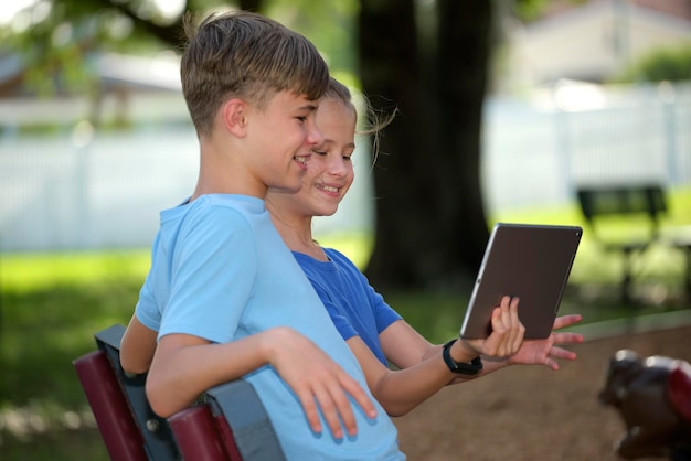 Dos niños felices, niña y niño, mirando la pantalla de la tableta digital leyendo estudiando o jugando sentados en un banco al aire libre en un día soleado de verano