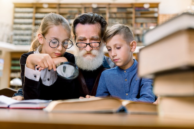Dos niños felices, niño y niña con lupa escuchando la interesante historia del libro de su guapo abuelo barbudo o maestro de escuela, sentados juntos en la antigua biblioteca.
