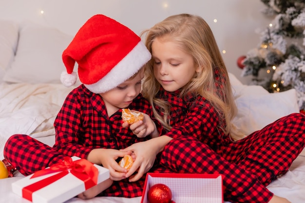 dos niños felices en pijama para Navidad comen mandarinas y juegan con juguetes navideños