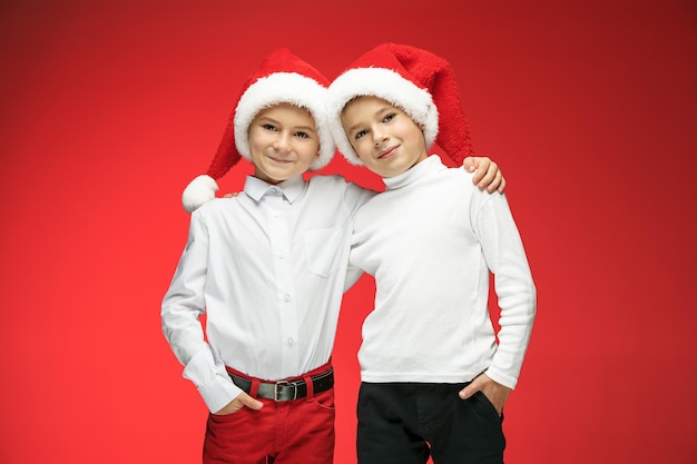 Dos niños felices con sombreros de santa claus con cajas de regalo en estudio rojo