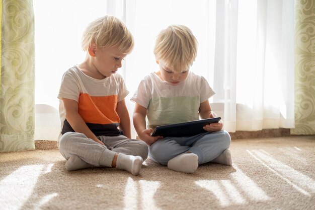 Dos niños gemelos divertidos usando tabletas viendo dibujos animados tirados en el suelo en casa