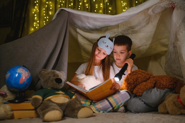 Dos niños con linterna leen un libro debajo de una manta a modo de carpa