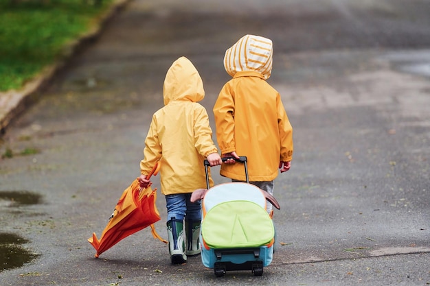 Dos niños con maleta de paraguas y capas impermeables amarillas y botas caminando juntos al aire libre después de la lluvia