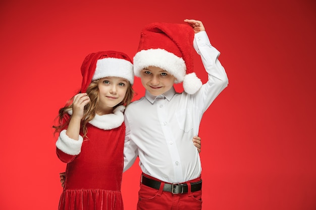 Dos niños y niñas felices con sombreros de santa claus con cajas de regalo en estudio rojo