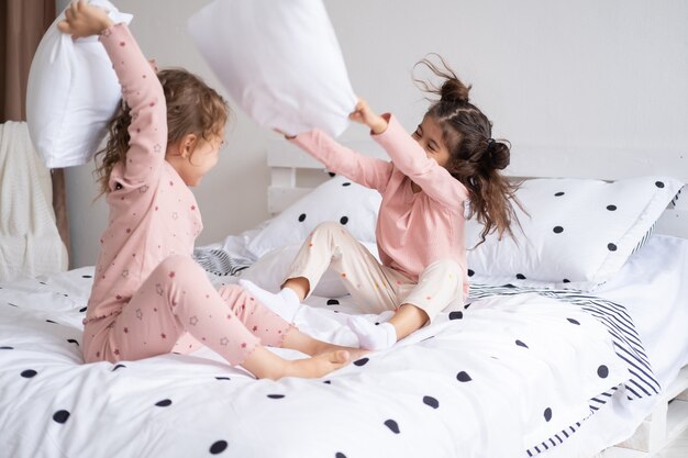 Dos niños niñas en pijama con pelea de almohadas en la cama en un apartamento moderno y luminoso