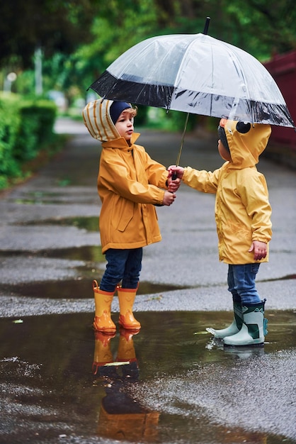Dos niños con paraguas en capas impermeables amarillas y botas jugando al aire libre después de la lluvia juntos