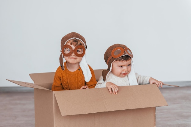 Dos niños pequeños con disfraces de piloto retro se divierten y se sientan en una caja de papel en el interior durante el día