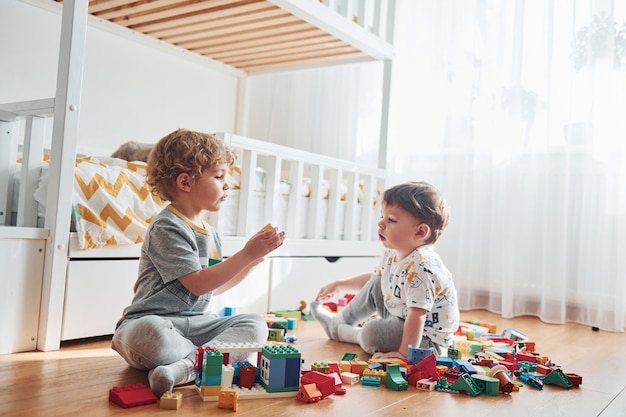 Dos niños pequeños se divierten en el interior del dormitorio con un juego de construcción de plástico El gato se sienta detrás de ellos