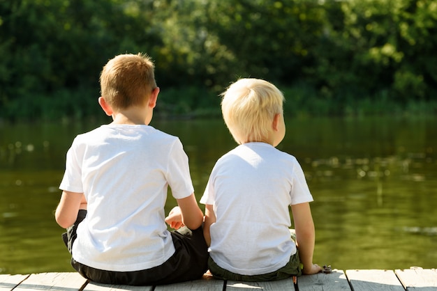 Dos niños pequeños están sentados en el muelle en la orilla del río. Concepto de amistad y fraternidad. Vista trasera