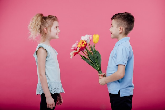 Dos niños pequeños. niño extiende flores a una niña en vestido rosa, aislado en la pared azul, vista de perfil