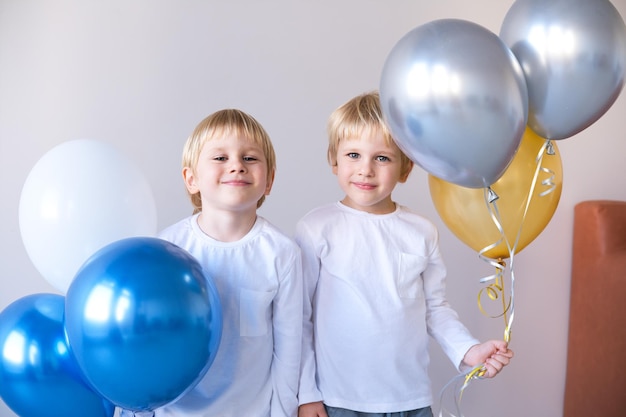 dos niños pequeños rubios sonrientes gemelos abrazados sosteniendo globos celebración de cumpleaños