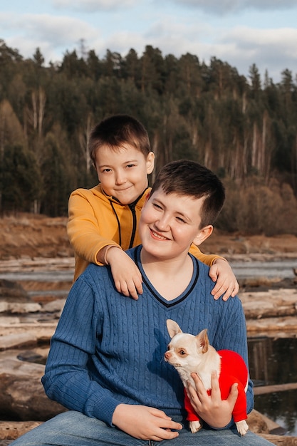 Dos niños pequeños se sientan en un abrazo sobre troncos junto al río en brazos de un perro chihuahua. El concepto de amistad y hermandad. Recreación al aire libre.