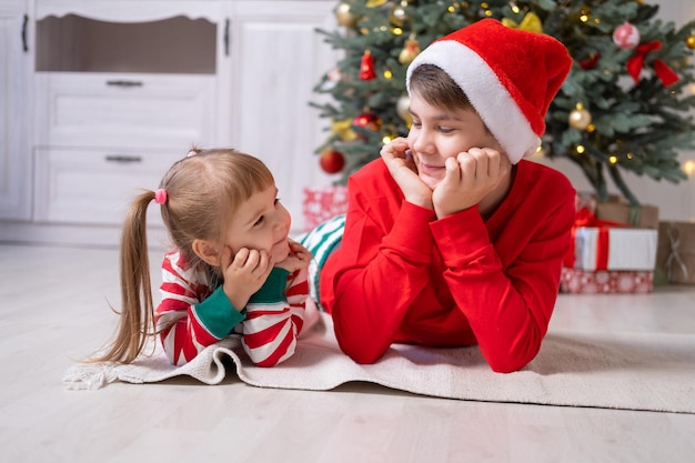 dos niños en pijama de Navidad con cajas de regalo bajo el árbol de Navidad en casa