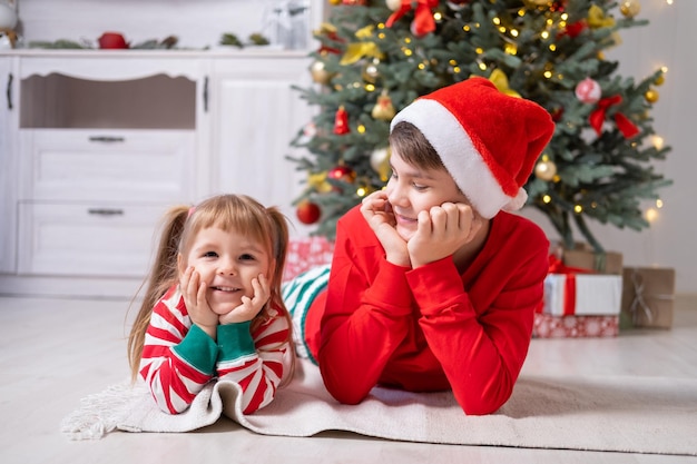 dos niños en pijama de Navidad con cajas de regalo bajo el árbol de Navidad en casa