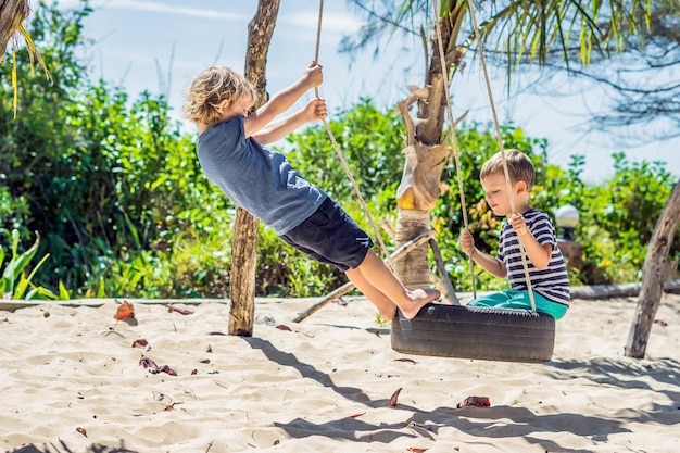 Dos niños rubios divirtiéndose en el columpio en la costa de arena tropical