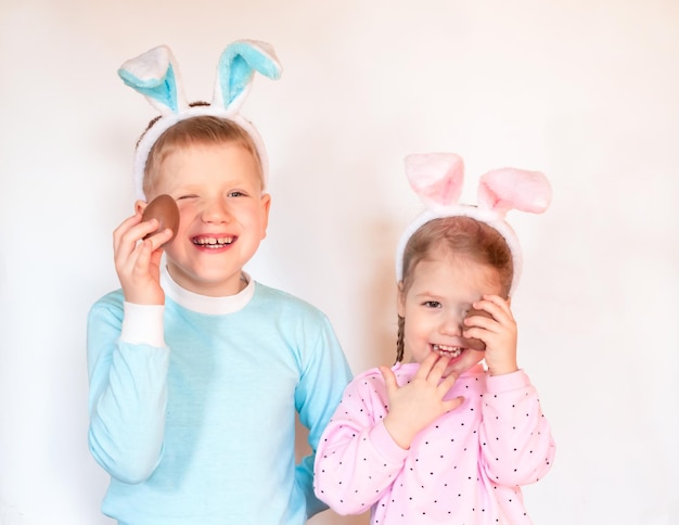 dos niños sonrientes con orejas de conejo de Pascua y huevos de chocolate sosteniendo frente a las caras