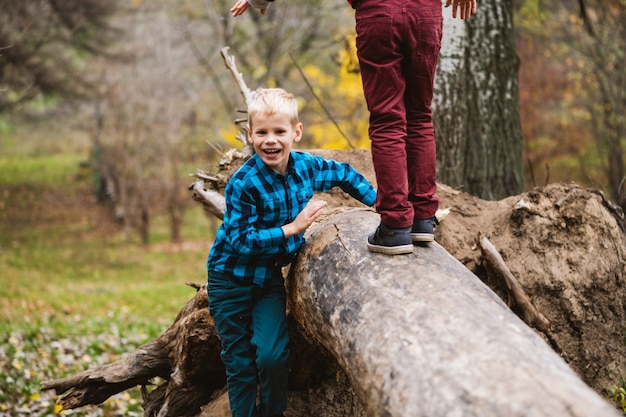 Dos niños varones preadolescentes curiosos trepando y jugando con un enorme tronco de árbol caído en el parque de otoño, infancia feliz activa