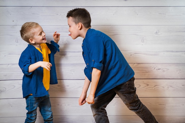 Dos niños vestidos de forma casual con fondo blanco de madera mostrándose lenguas el uno al otro Dos hermanos jugando Hermanos mayores y hermanos menores Concepto de infancia feliz Amistad