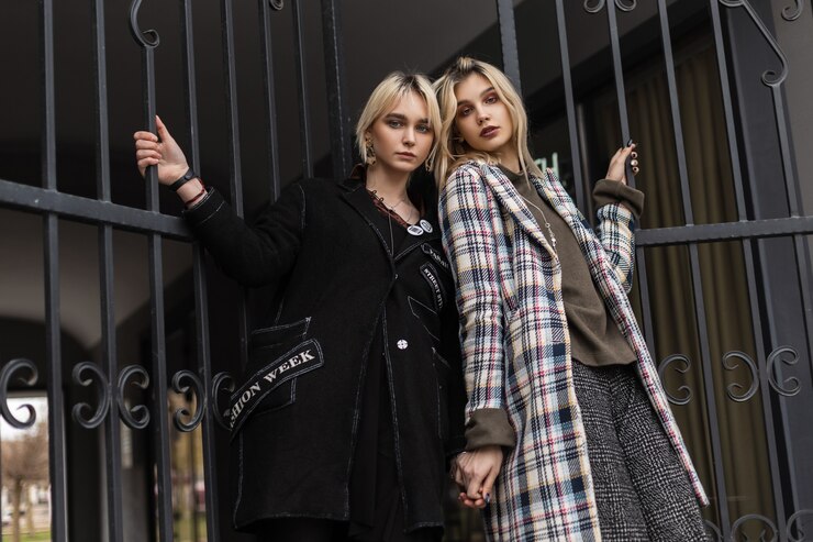 Foto dos novias rubias jóvenes de moda en chaquetas elegantes de gran tamaño posando cerca de la puerta de hierro en la calle