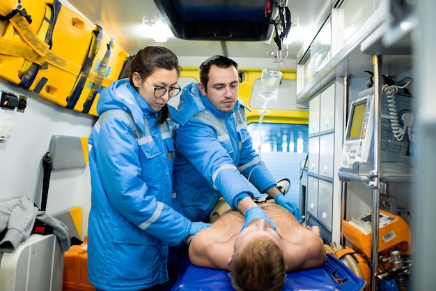 Uno de los dos paramédicos que controlan el pulso del hombre sin camisa inconsciente en camilla con un colega de pie cerca