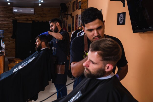 Foto dos peluqueros barbudos dando cortes de pelo a clientes masculinos en la barbería - barberos trabajando en su salón de hombres.