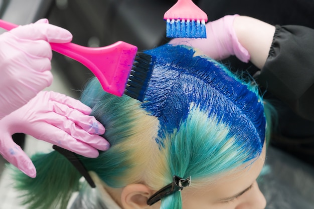 Dos peluqueros usando un pincel rosa mientras aplican pintura azul a una clienta con cabello color esmeralda ...