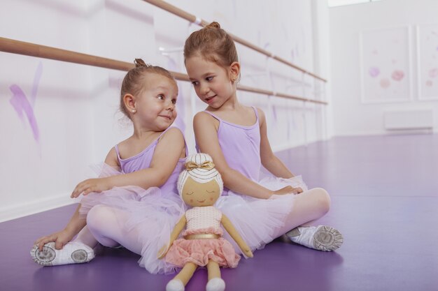 Dos pequeñas bailarinas lindas sentadas espalda con espalda en el suelo, mirándose sobre sus hombros