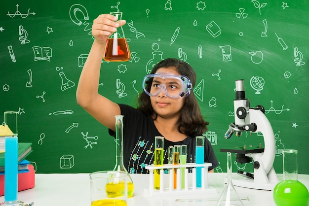 Dos pequeñas y lindas colegialas indias o asiáticas experimentando o estudiando ciencias en laboratorio, sobre fondo de pizarra verde con garabatos educativos